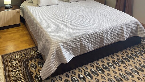 A neatly made bed with a white quilt, two bedside lamps, a framed artwork on the wall, and a patterned rug on the floor. - Dara House (Plovdiv)