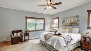 A bedroom with a large bed, a ceiling fan, a window with blinds, a chair, a dresser, and a painting on the wall.