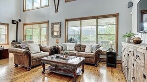 A spacious living room with two leather sofas, a wooden coffee table, and large windows offering a view of greenery.