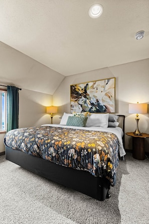 A bedroom with a large bed, two bedside lamps, a ceiling light, a window with curtains, and a painting on the wall.