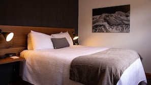 Una cama bien hecha con cabecera de madera, dos lámparas de noche y un cuadro de un paisaje montañoso en la pared.