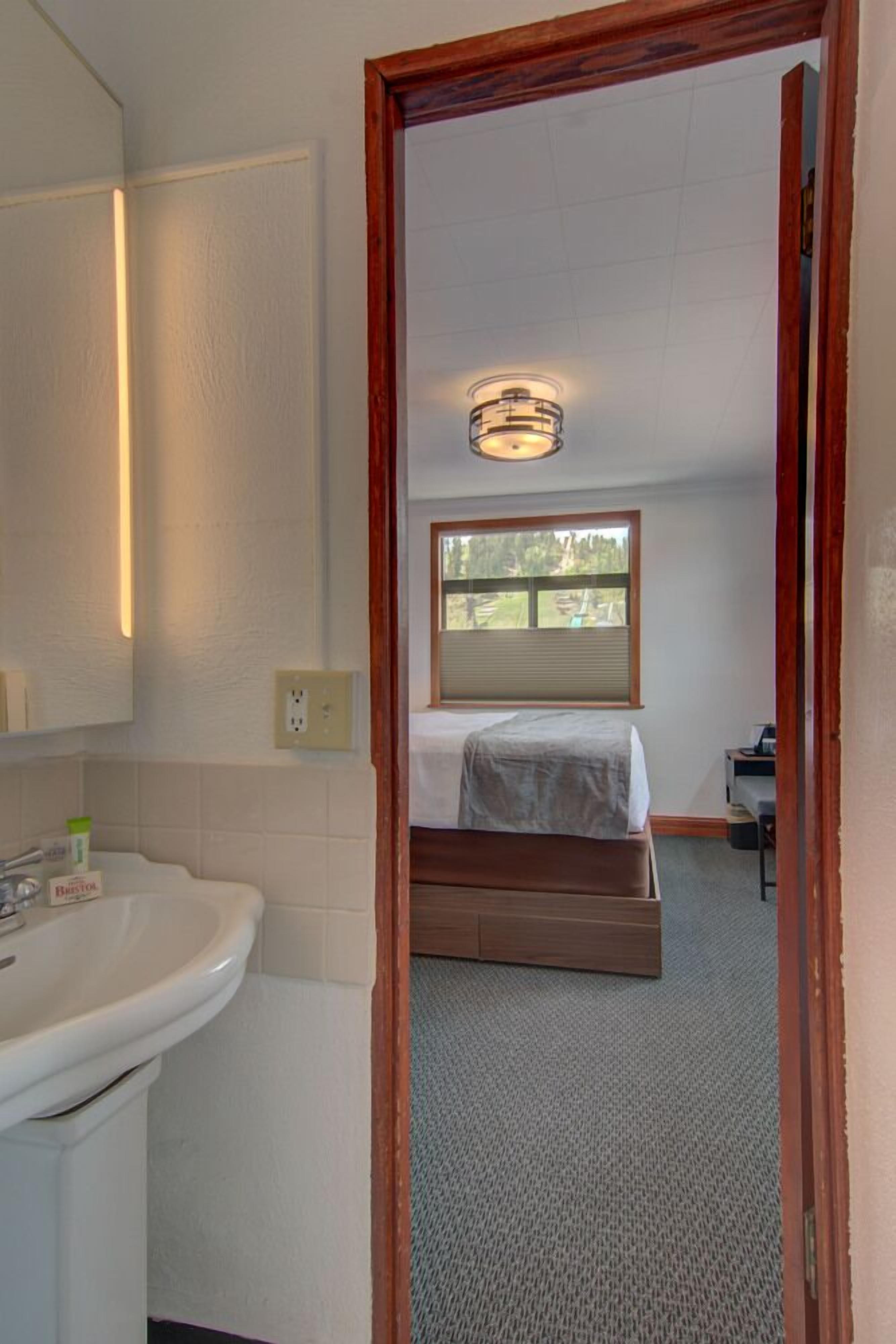A bedroom with a bed, a window, a bedside table, and a bathroom with a sink and mirror.