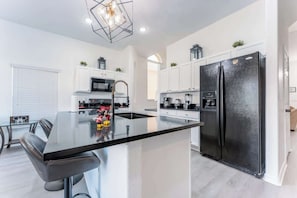 A modern kitchen with a central island, white cabinetry, a black refrigerator, and a dining area with a glass table and chairs. - Luxury Family Villa w Private Pool 10 min Disney (Davenport)