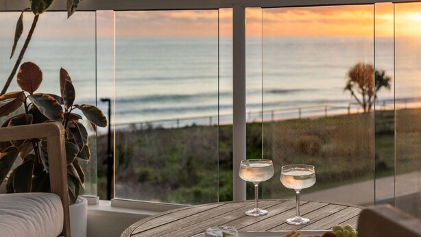 A balcony with a view of the ocean at sunset, a round wooden table with two glasses of water, a tray with snacks, and a potted plant. - 2 Brm Beachfront Retreat ~ Azure Beachfront Bliss (Palm Beach)