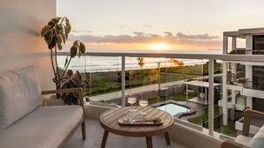 A balcony with a sofa, a wooden table with glasses and snacks, and a view of a swimming pool and landscape at sunset. - 2 Brm Beachfront Retreat ~ Azure Beachfront Bliss (Palm Beach)
