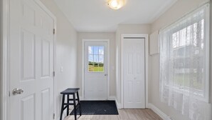 Un pasillo con puertas blancas, una mesita pequeña, un felpudo y una ventana con cortinas de encaje.