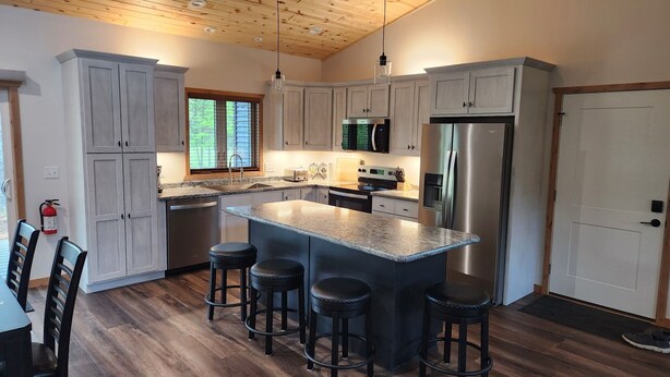 A modern kitchen with grey cabinets, a central island with stools, stainless steel appliances, and a wooden ceiling.
