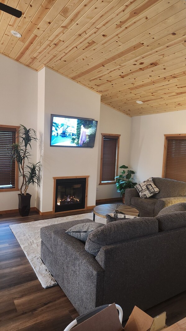 A living room with a wooden ceiling, a flat-screen TV mounted on the wall, a fireplace, and a large sectional sofa. - New 4-bdrm house Sand Valley/Lake Arrowhead area. 5 min from world class golf. (Nekoosa)