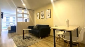 A modern living room with a sofa, a small table, and a chair. There are framed pictures on the wall and a window with blinds.