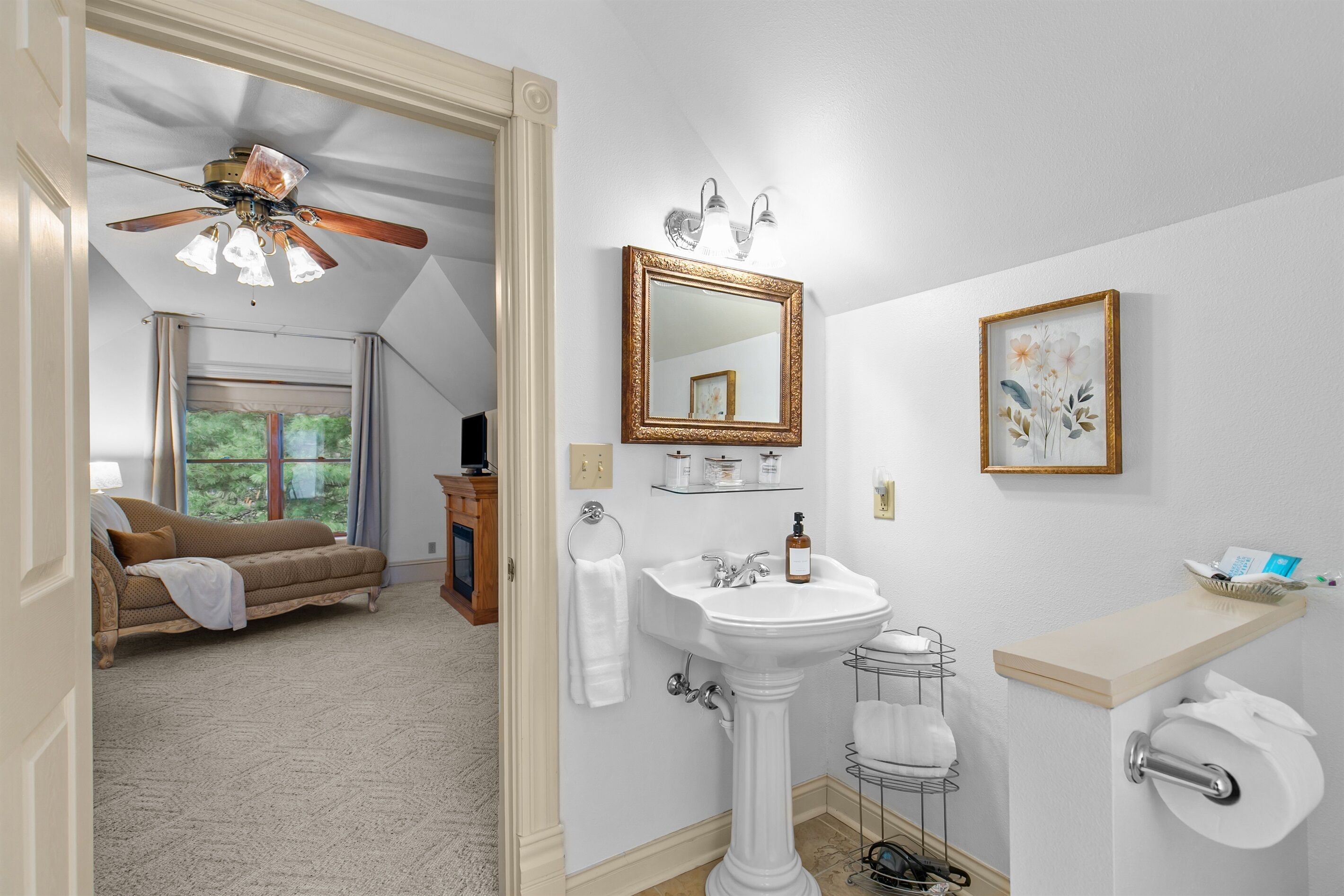 A bathroom with a pedestal sink, a mirror, a ceiling fan, a toilet, and a window with curtains.