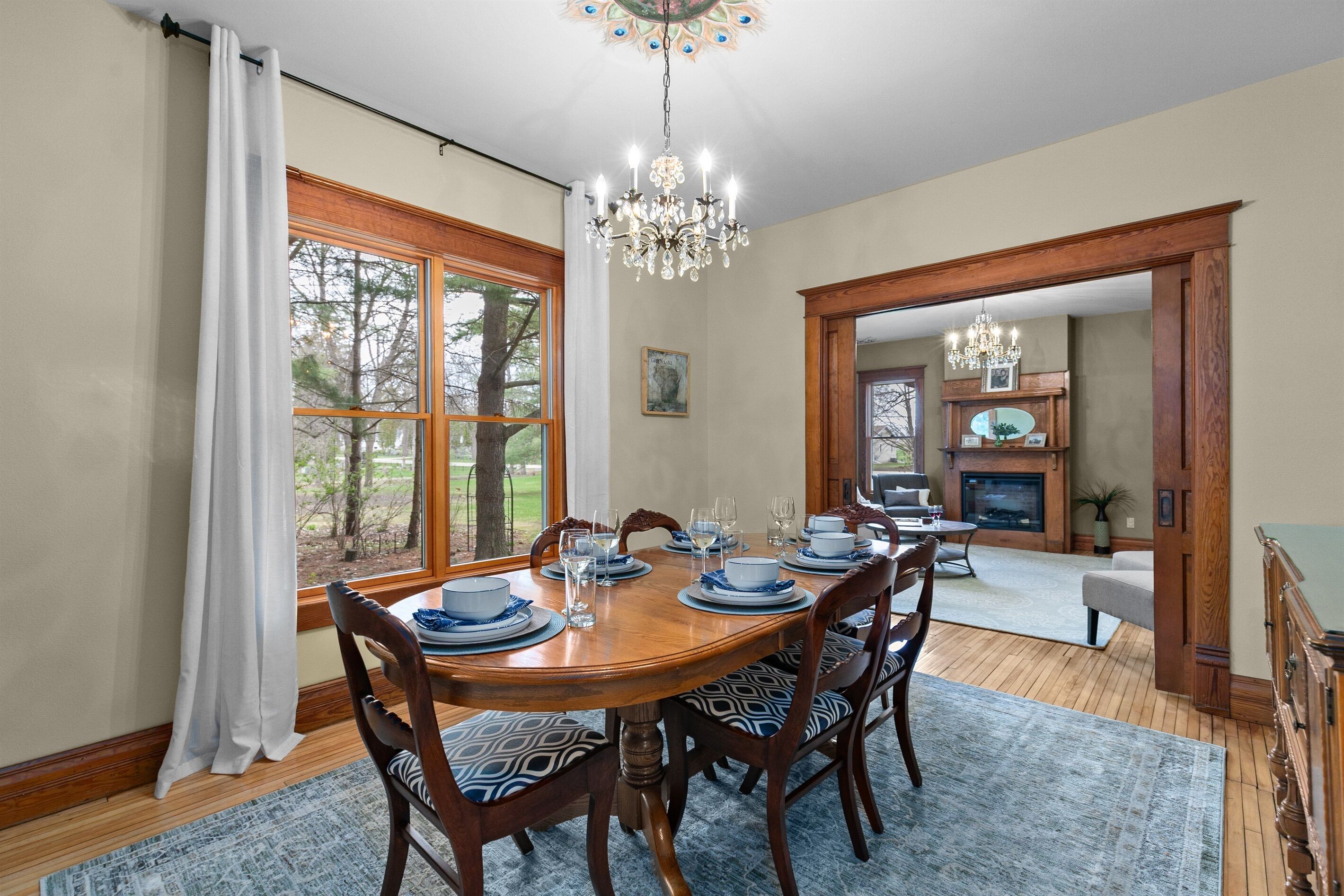 A dining room with a wooden table set for six, a chandelier, and a view through a window.