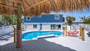 A pool area with a thatched roof, a pool, and red lounge chairs.