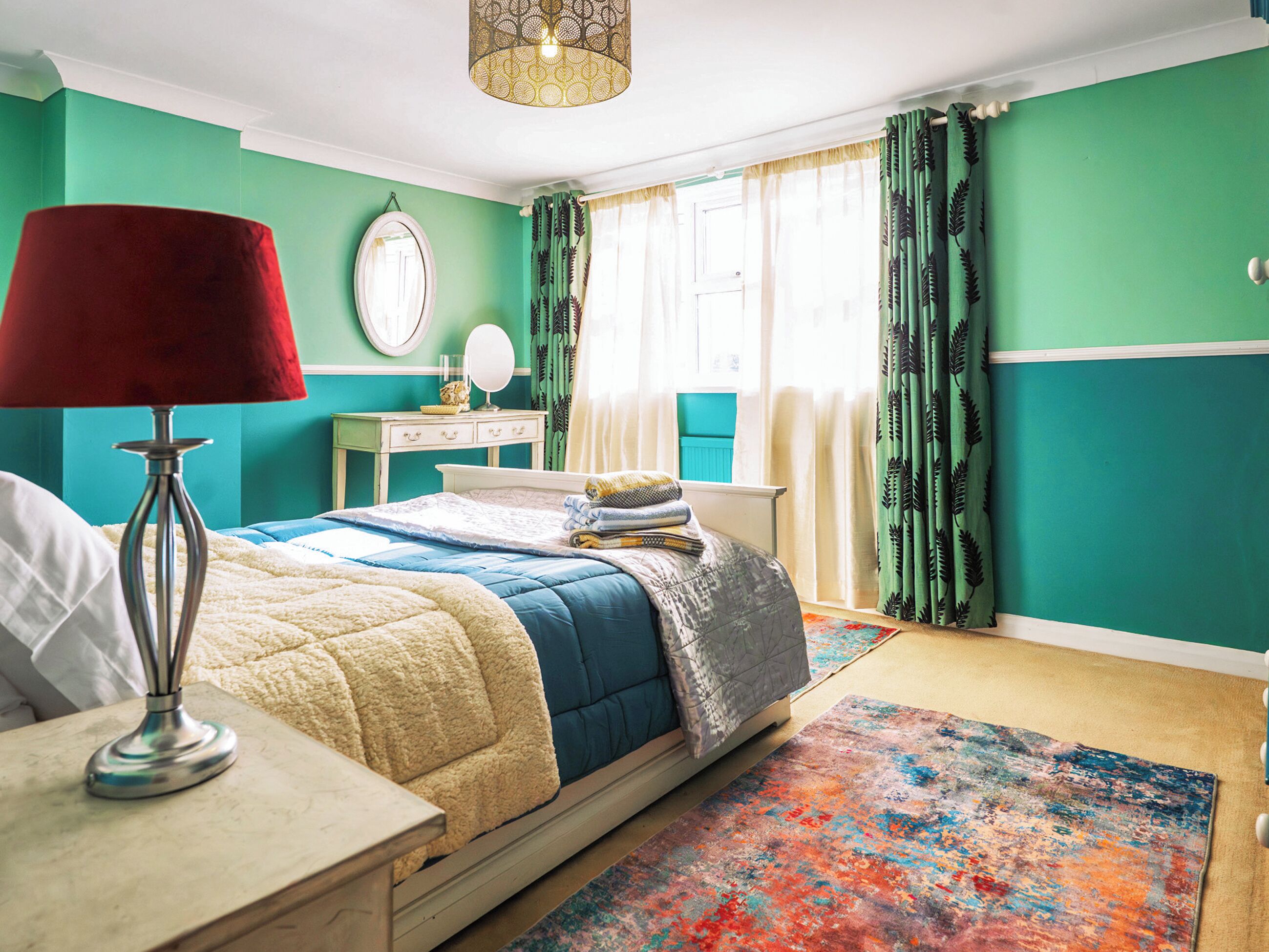 A bedroom with a bed, bedside table, lamp, dresser, mirror, and a window with curtains.