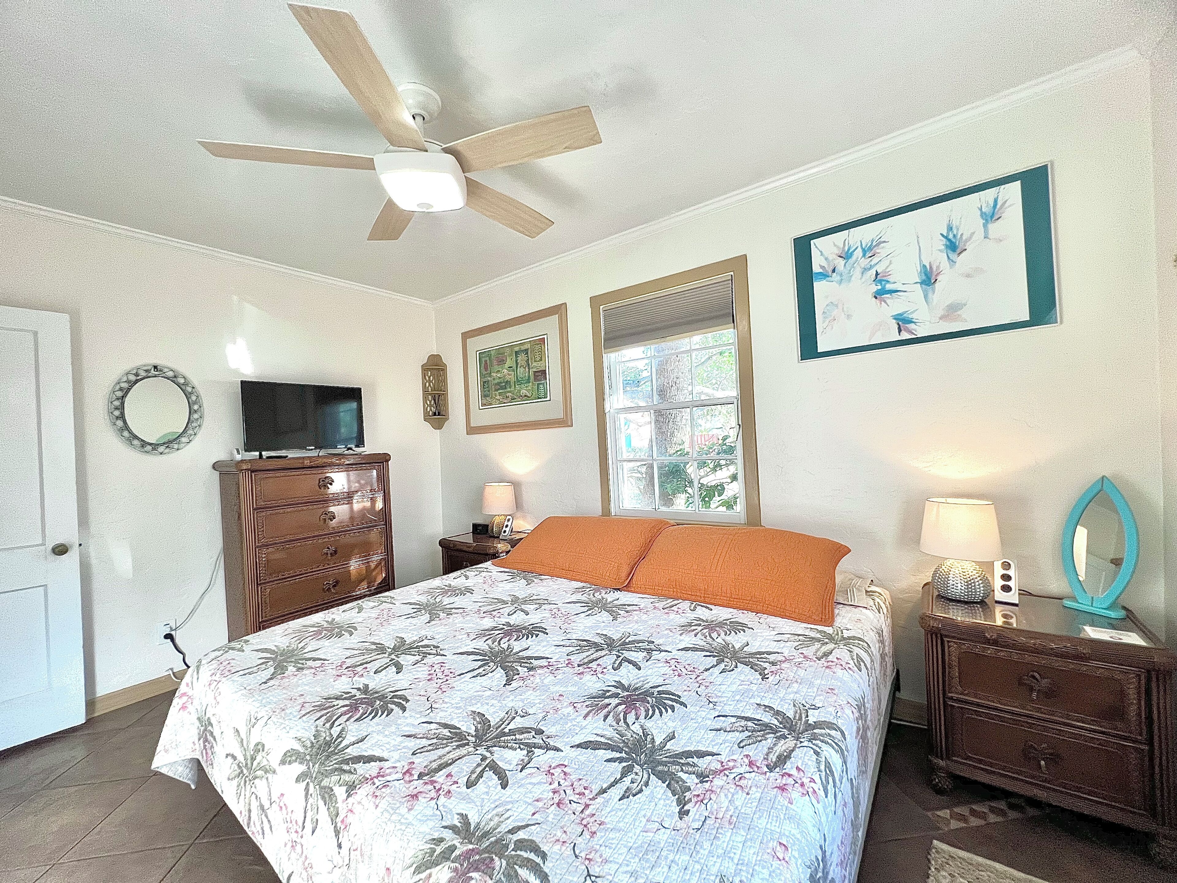 Une chambre à coucher comprenant un lit, un ventilateur de plafond, une commode, un téléviseur et une fenêtre avec des stores.