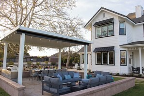 A covered patio with outdoor furniture and a fire pit, adjacent to a two-story house with large windows and a covered entryway.