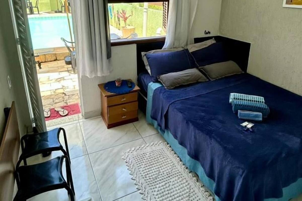 A bedroom with a bed, a small table, a chair, a window with curtains, and a view of a swimming pool.