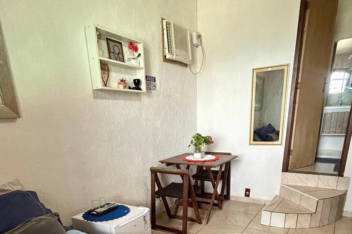 A hotel room with a bed, a small table with a chair, a mirror, and a shelf with decorative items.