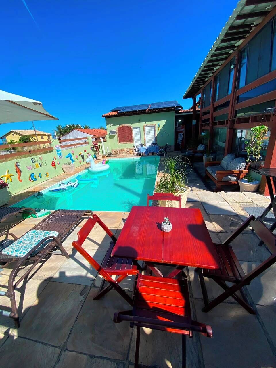 Zona de piscina con sillas de descanso y una mesa, rodeada por un edificio de fachada verde y un cielo azul despejado.