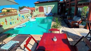 Zona de piscina con sillas de descanso y una mesa, rodeada por un edificio de fachada verde y un cielo azul despejado.