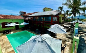 A pool area with umbrellas, a pool, and a building with a tiled roof. - Pousada Aconchego de Maricá (Marica)