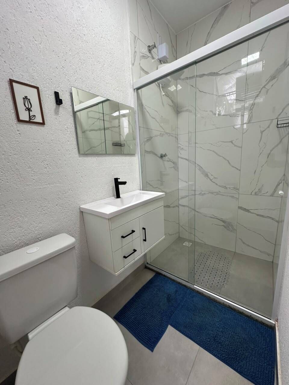Een badkamer met een wit toilet, een witte wastafel met een zwarte kraan, een glazen douchecabine en een blauw vloerkleed.