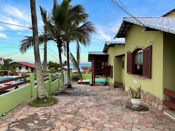 Zona residencial con un camino pavimentado, palmeras y una casa con techo de tejas y contraventanas de madera.