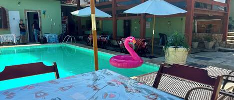 Ein Poolbereich mit einem Tisch, der mit einer Tischdecke mit Kaffeemotiven bedeckt ist, einem rosa Flamingo-Schwimmtiere und Sonnenschirmen.