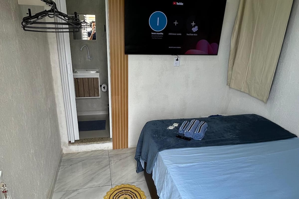 A bedroom with a bed, a TV mounted on the wall, a towel rack, and a small mat on the floor.