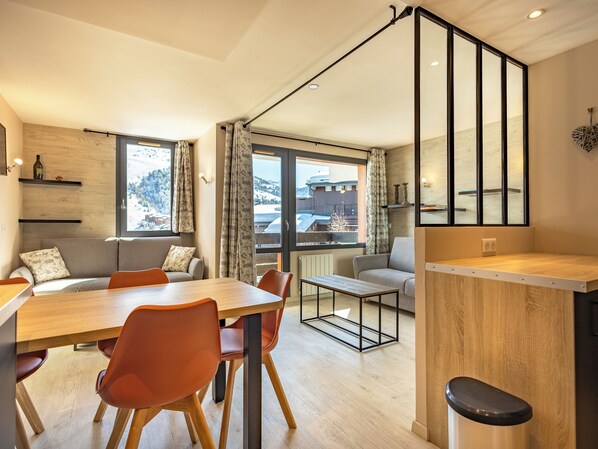 Une cuisine moderne avec une table à manger en bois, des chaises orange et une vue sur des montagnes enneigées, visible à travers de grandes fenêtres.