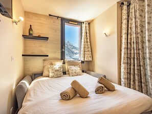 Une chambre avec un grand lit, une tête de lit en bois, une bouteille posée sur une étagère et, par la fenêtre, une vue sur des montagnes enneigées.