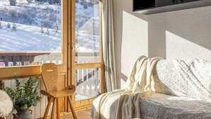 A cozy room with a bed, a wooden chair, a potted plant, and a view of a snowy landscape through a large window.