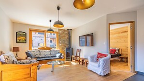 A spacious living area with wooden furniture, a fireplace, and a large window providing a view of the outside. - Chalet Olympe #101 - 5 guests (Les Allues)