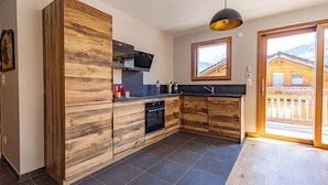A modern kitchen with wooden cabinetry, a built-in oven, and a large window offering a view of a wooden cabin. - Chalet Olympe #101 - 5 guests (Les Allues)