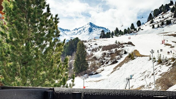 Una terraza de madera con vistas a un paisaje montañoso nevado, que incluye una pista de esquí y árboles.