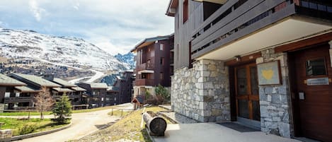 Un edificio moderno con fachada de piedra y madera, una entrada de piedra y vistas a montañas nevadas al fondo.