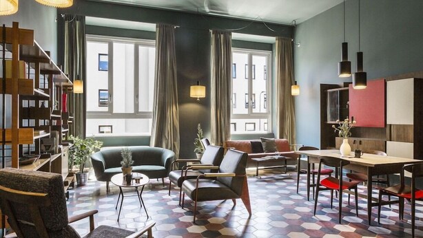 Een moderne woonkamer met een zwart-wit geschilderd vloerkleed, een bank, fauteuils, een eettafel met stoelen, een boekenkast en grote ramen voorzien van gordijnen.