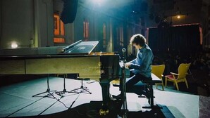 Una persona tocando el piano sobre un escenario, con un soporte de micrófono, un piano y algunas sillas al fondo.