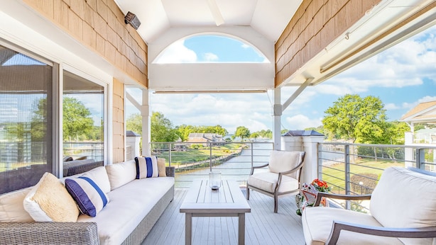 Un espace extérieur abrité comprenant un canapé, des fauteuils et une table. Il est doté de grandes fenêtres et offre une vue sur une rivière et la verdure environnante.