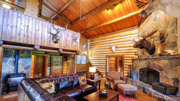 A log cabin interior with a stone fireplace, leather furniture, and mounted animal heads.