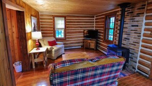 A log cabin interior with a sofa, a table with a lamp, a television, and a fireplace. - Historic Pioneer Cabin (Coram)