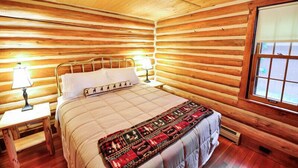 A log cabin room with a bed, a nightstand, a lamp, and a window. - Historic Pioneer Cabin (Coram)