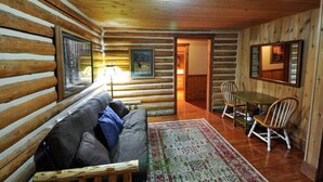 A log cabin interior with a wooden floor, a sofa, a dining table with chairs, and a large window. - Historic Pioneer Cabin (Coram)