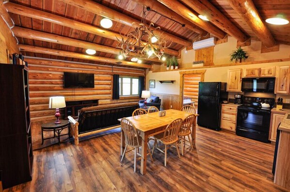 A log cabin interior with a wooden dining table, chairs, a fireplace, a kitchen with wooden cabinets, and a ceiling with exposed beams.
