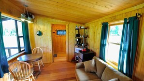 A wooden cabin interior with a wooden dining table, two chairs, a sofa, a kitchen area with shelves, and large windows with curtains.