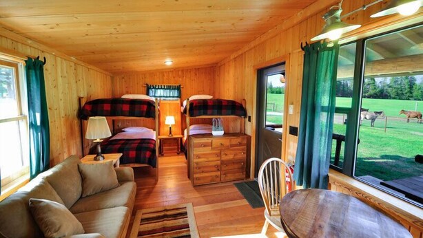 A cozy cabin interior with two bunk beds, a sofa, a wooden table, and a large window offering a view of a grassy field.