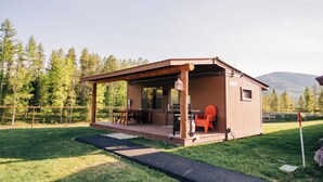 A small wooden cabin with a covered porch, a table, and chairs, surrounded by a grassy yard and trees.
