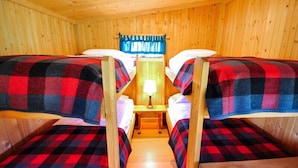 A cabin with two bunk beds, each with a plaid blanket, a small table with a lamp, and a window with blue curtains.