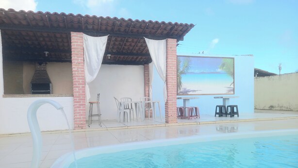 Uma piscina com um deck ladrilhado, um espaço coberto com terraço e cadeiras, e uma parede de tijolos.