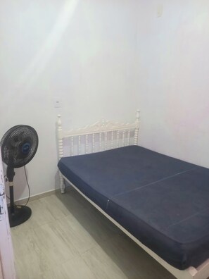 Ein Einzelbett mit weißem Kopfteil und dunkler Matratze, ein Standventilator und ein heller Holzboden.