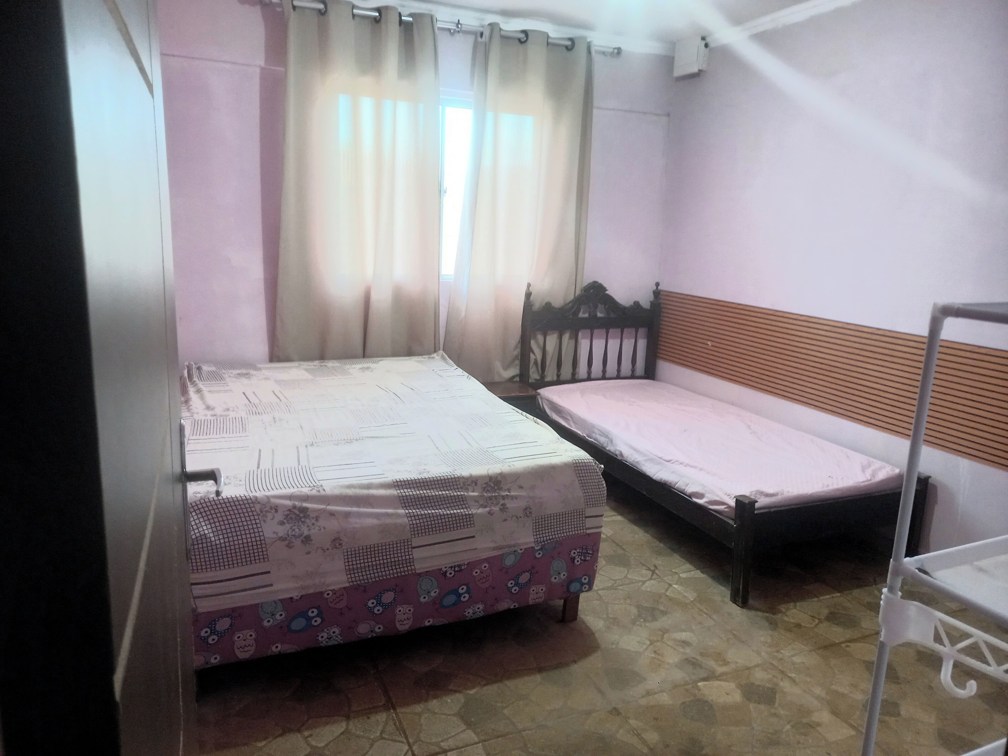 Quarto com duas camas, janela com cortinas, chão em azulejo e prateleira de metal.